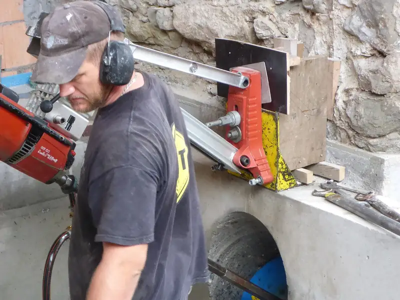 Worker in hearing protection and gloves running a wall-mounted diamond core drill beside a freshly cored concrete opening